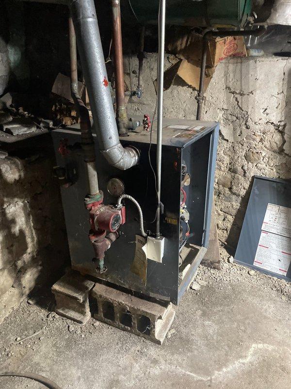 Conducted comprehensive assessment of client's aging LP boiler hot water heating system. Observed original installation mounted on concrete blocks with connected galvanized ductwork, copper piping, and red circulator pump showing significant wear and corrosion. System remains operational but has reached end of service life; documented deteriorating components and electrical connections for replacement estimate.